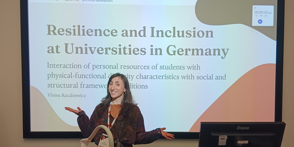 Auf dem Bild steht eine Person an einem Rednerpult in einem Vortragsraum und hält eine Präsentation. Hinter ihr ist eine große Präsentationsfolie zu sehen mit dem Titel „Resilience and Inclusion at Universities in Germany“. Auf der Folie sind außerdem Logos der TU Dortmund zu erkennen.