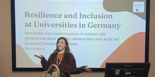 Auf dem Bild steht eine Person an einem Rednerpult in einem Vortragsraum und hält eine Präsentation. Hinter ihr ist eine große Präsentationsfolie zu sehen mit dem Titel „Resilience and Inclusion at Universities in Germany“. Auf der Folie sind außerdem Logos der TU Dortmund zu erkennen.