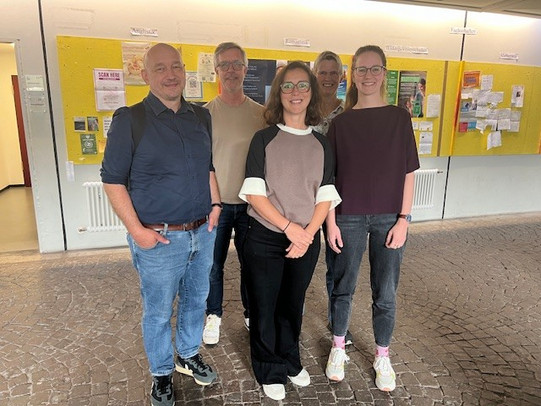 In einer modernen Umgebung stehen fünf Personen zusammen und posieren für ein Foto. Sie tragen legere Kleidung, die ihren individuellen Stil widerspiegelt. Im Hintergrund sind Informationswände mit verschiedenen Aushängen zu sehen, was auf eine lebendige und aktive Atmosphäre hinweist. Die Gruppe wirkt freundlich und entspannt, während sie diesen Moment festhält.