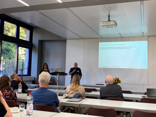 Bei einer Veranstaltung an der TU Dortmund präsentieren zwei Referenten vor einem interessierten Publikum. Im Hintergrund ist eine Präsentation mit dem Thema "Bildung und Schule" zu sehen, die verschiedene Aspekte wie Didaktik, Grundsätze und Fördermöglichkeiten behandelt. Die Anwesenden hören aufmerksam zu, während die Referenten ihre Ideen und Konzepte vorstellen. Die Atmosphäre im Raum ist entspannt und förderlich für den Austausch von Wissen und Erfahrungen.