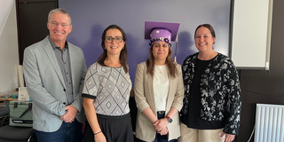 Dir. U. Prof. Dr. Lars Adolph (baua), Prof. Dr. Liane Bächler (TU Dortmund), Sara Kerdar (baua), Dr. Britta Kirchhoff (baua) Vier Personen stehen nebeneinander in einem Seminarraum und lächeln in die Kamera. Im Hintergrund ist ein großer Bildschirm mit einer darauf montierten Webcam sowie ein Whiteboard zu sehen. Von links: ein Mann mit grauen Haaren und Brille in einem hellgrauen Blazer und dunkler Jeans; eine Frau mit dunklen Haaren und runder Brille in einem weiß-schwarz gemusterten Oberteil und schwarzer Hose; eine Frau mit einem lila, blumenverzierten Doktorhut, die einen hellen Blazer über einem weißen Oberteil trägt; und eine Frau mit zurückgebundenen dunklen Haaren in einer schwarzen Bluse mit weißem Blumenmuster und heller Hose.