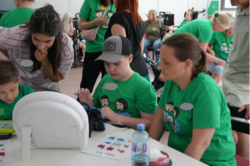 Mehrere Menschen befinden sich in einem Raum und arbeiten gemeinsam an einem Tisch. Im Vordergrund sitzen zwei Personen in grünen T‑Shirts neben einer weiteren Person und nutzen ein Tablet mit Halterung. Auf dem Tisch liegen Karten mit Symbolen sowie eine Wasserflasche. Im Hintergrund sind weitere Personen zu sehen, einige tragen ebenfalls grüne T‑Shirts und einige nutzen Rollstühle oder andere Hilfsmittel.