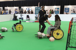 Foto. Zwei Personen im Rollstuhl spielen mithilfe eines an dem Rollstuhl befestigten Geräts Fußball.