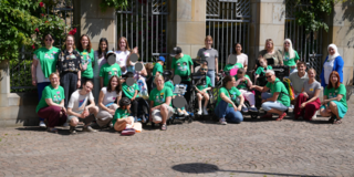 Eine größere Gruppe von Menschen posiert im Freien für ein Gruppenfoto vor einem Gebäude mit steinernen Bögen, Metallgittern und Kletterpflanzen. Viele tragen grüne T‑Shirts mit einem Aufdruck. Einige Personen sitzen oder knien vorne, andere stehen dahinter. Mehrere Teilnehmende nutzen Rollstühle oder andere Hilfsmittel. Der Platz vor ihnen ist mit Kopfsteinpflaster bedeckt, und es ist sonniges Wetter. 