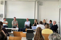 Ein Seminarraum mit mehreren Zuhörenden, die an Tischen sitzen und nach vorne schauen. Vorne sitzen mehrere Personen in einer Reihe und diskutieren im Rahmen einer Podiumsrunde. Hinter ihnen befindet sich eine große weiße Projektionsfläche und darunter eine grüne Tafel. Einige der Zuhörenden haben Laptops vor sich auf den Tischen. Die Situation wirkt wie eine moderierte Diskussion oder ein Panel im Rahmen einer universitären Veranstaltung.