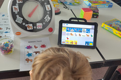 Ein Kind sitzt an einem Tisch und blickt auf ein Tablet mit einer Symboloberfläche. Daneben steht ein großes Drehrad mit Bildern von Tieren. Auf dem Tisch liegen außerdem ein Bingo‑Spielblatt mit Tiermotiven, ein Glas mit kleinen Sternen sowie mehrere Spiele und Materialien. Hinter dem Tisch stehen Stühle in einem Raum.