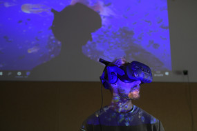Im Vordergrund ein Mann mit Kinnbart, der ein VR-Headset trägt. Hinter ihm eine bläuliche Projektion mit Quallen und anderen Meerestieren. Ein Ausschnitt des Bildes wird vom Schatten seines Kopfes verdeckt, das Gesicht ist blau erleuchtet.