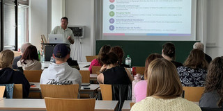 Ein Seminarraum mit mehreren Studierenden, die an Tischen sitzen und nach vorne schauen. Vorne steht eine Person neben einem Rednerpult mit Laptop und hält einen Vortrag. Auf einer Leinwand wird eine Präsentationsfolie mit der Überschrift „Was wir gehört haben“ gezeigt, darunter mehrere Stichpunkte. Hinter der Leinwand befindet sich eine grüne Tafel. Links im Raum sind große Fenster, durch die Tageslicht hereinfällt. Die Szene wirkt wie eine Vorlesung oder Präsentation an einer Universität.