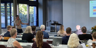 Ein engagierter Redner steht vor einer aufmerksamen Zuhörerschaft in einem modernen Seminarraum der TU Dortmund. Die Teilnehmer, sowohl Studierende als auch Mitarbeitende, verfolgen gespannt die Präsentation, während im Hintergrund eine Leinwand mit Informationen und Grafiken zu sehen ist. Der Raum ist hell erleuchtet und fördert eine angenehme Lernatmosphäre. Auf den Tischen stehen Getränke und Notizmaterialien bereit, um das Lernen und den Austausch zu unterstützen.