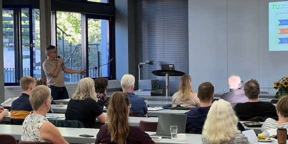 Ein engagierter Redner steht vor einer aufmerksamen Zuhörerschaft in einem modernen Seminarraum der TU Dortmund. Die Teilnehmer, sowohl Studierende als auch Mitarbeitende, verfolgen gespannt die Präsentation, während im Hintergrund eine Leinwand mit Informationen und Grafiken zu sehen ist. Der Raum ist hell erleuchtet und fördert eine angenehme Lernatmosphäre. Auf den Tischen stehen Getränke und Notizmaterialien bereit, um das Lernen und den Austausch zu unterstützen.