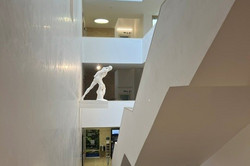 Beitrag ICCHP in Brno Blick in einen hellen, mehrstöckigen Innenhof (Atrium) eines modernen Gebäudes mit weißen Wänden und offenen Galerien. Auf einer mittleren Etage steht eine weiße Skulptur einer sich vorbeugenden Person. Unten im Foyer steht ein schwarzes Flügelklavier, rechts verläuft eine schräge, weiße architektonische Struktur (Treppe). Im Hintergrund unten sind Türen, ein Aufzug und ein Info-Punkt zu erkennen.