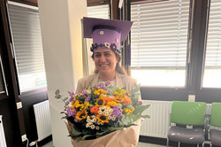 ine lächelnde Frau steht in einem hellen Raum mit Fenstern und grünen Stühlen im Hintergrund. Auf dem Kopf trägt sie einen mit Blumen verzierten lila Doktorhut, in den Händen hält sie einen großen, bunten Blumenstrauß mit Sonnenblumen, Chrysanthemen und Gänseblümchen. Sie trägt einen hellen Blazer, eine schwarze Hose und weiße Schuhe.