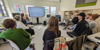 Foto. Zehn Menschen sitzen in einem Stuhlhalbkreis und schauen auf eine Präsentation.
