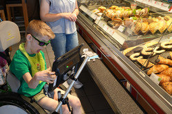 Ein Kind sitzt in einem Rollstuhl vor einer Bäckereitheke mit verschiedenen belegten Brötchen, Gebäck und Croissants hinter einer Glasscheibe. Vor dem Kind ist ein Tablet an einer Halterung befestigt, das es mit der Hand bedient. Neben dem Kind steht eine erwachsene Person.