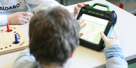 Foto. Kind von hinten, zeigt auf einen Talker. Daneben ein Brettspiel mit zwei Figuren