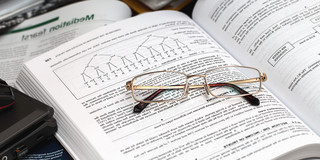 Auf dem Bild liegt eine aufgeschlagene Fachbuchseite mit Diagrammen und Texten, auf der ein Paar goldfarbener Brillen platziert ist. Rundherum befinden sich weitere Bücher, Unterlagen und ein Laptop, was auf eine Lern- oder Arbeitssituation hindeutet. Das Bild vermittelt den Eindruck konzentrierten Studierens oder wissenschaftlicher Recherche.