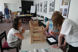 Mehrere Personen befinden sich in einem Raum mit Tischen und Stühlen und arbeiten gemeinsam an einer Aktivität. Im Vordergrund stehen drei Personen an einem Tisch mit einer Holzkiste, verschiedenen Materialien und einem Tablet mit Symboloberfläche. Eine Person hält einen Gegenstand in der Hand, während die anderen zuschauen oder unterstützen. An den Wänden hängen gerahmte Bilder. Im Hintergrund sind weitere Personen und Spielmaterialien zu sehen.