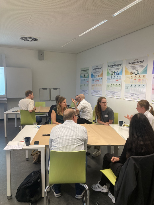 Teilnehmende sitzen in Kleingruppen an Tischen mit Papiertischdecken in einem Seminarraum und diskutieren; an der Wand hängen mehrere Workshop-Poster mit Leitfragen (u. a. „Muss das wirklich ins Studium?", „Reicht Sensibilisierung?", „Wer ist eigentlich zuständig?", „Was kann dafür weg?") und Abstimmungsrastern.