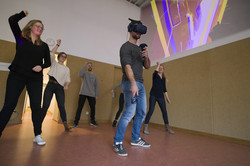 Teile des Projektteams und Studierende im Bewegungsambulatorium, anscheinend tanzend; ein Mann steht mit VR-Headset und Handcontrollern in der Mitte des Raumes, hinter ihm eine bunte Projektion.