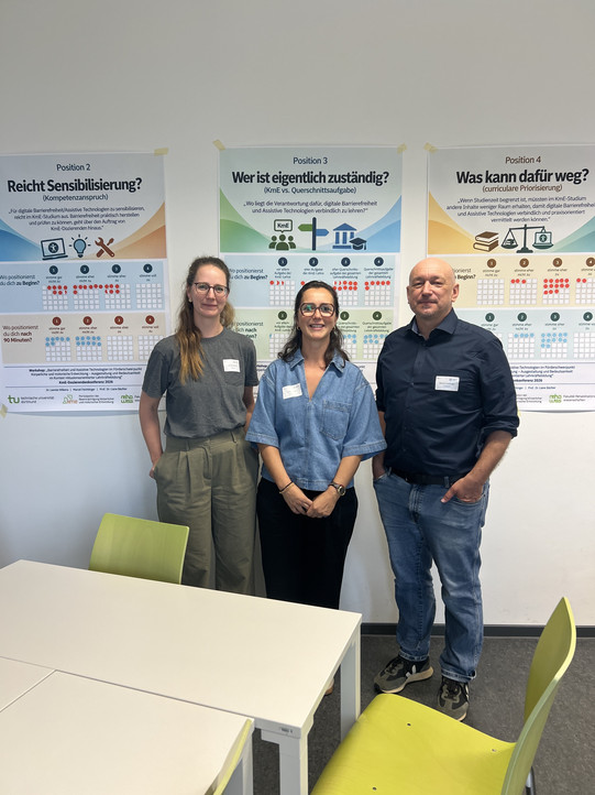 Drei Personen (zwei Frauen und ein Mann) mit Namensschildern stehen nebeneinander und lächeln in die Kamera, vor einer Wand mit drei Workshop-Postern zu den Themen „Reicht Sensibilisierung?", „Wer ist eigentlich zuständig?" und „Was kann dafür weg?"; im Vordergrund sind ein weißer Tisch und grüne Stühle zu sehen.