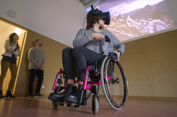 Eine Frau im Rollstuhl in Mitten des Bewegungsambulatoriums. Sie trägt VR-Ausrüstung. Im Hintergrund zwei Personen, die auf eine Projektion an der Wand schauen; die Projektion zeigt eine Berglandschaft.