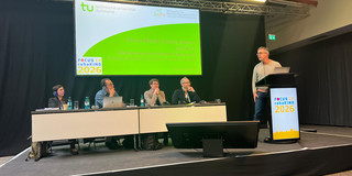 Foto. Riclef Schomerus an einem Rednerpult auf einer Bühne. Hinter ihm sitzen vier Menschen, die ihn anschauen. Auf der Präsentationsfolie im Hintergrund steht: "Gross Motor Family Report (GM-FR). Übersetzung und kulturelle Anpassung für Deutschland, Österreich und die Schweiz"