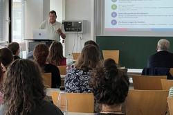 Ein Seminarraum an einer Universität. Vorne steht eine Person an einem Rednerpult mit Laptop und hält einen Vortrag. Hinter ihr wird eine Präsentationsfolie mit dem Titel „Was wir gehört haben“ auf eine Leinwand projiziert. Darauf sind mehrere Stichpunkte aufgelistet. Im Raum sitzen zahlreiche Zuhörerinnen und Zuhörer auf Stühlen an Tischen und schauen nach vorne zur Präsentation. Links sind große Fenster, die Tageslicht in den Raum lassen, und vorne befindet sich eine grüne Tafel unter der Leinwand. Die Atmosphäre wirkt wie eine Vorlesung oder ein wissenschaftlicher Vortrag.