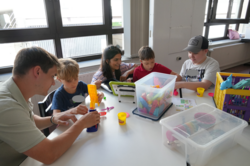 Mehrere Kinder sitzen gemeinsam mit einer erwachsenen Person an einem Tisch in einem hellen Raum mit großen Fenstern. Auf dem Tisch liegen verschiedene Spielmaterialien, darunter bunte Knete, Formen und Werkzeuge. Die Kinder beschäftigen sich mit dem Formen und Basteln der Knete, während die erwachsene Person sie dabei unterstützt.