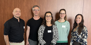 Fünf Personen (zwei Männer und drei Frauen), teils mit Namensschildern, stehen nebeneinander vor einer braunen Wand und lächeln in die Kamera.