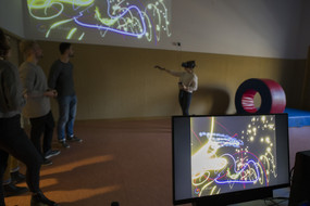 Rechts im Vordergrund ein PC-Bildschirm, darauf bunte Lichtstreifen. Im Hintergrund einige Personen, eine davon mit VR-Equipment; sie streckt ihren Arm in die Raummitte, um so in der virtuellen Realität mit den Lichtstreifen zu zeichnen.