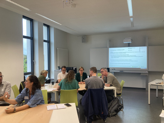 Teilnehmende sitzen in Kleingruppen an mit Papier bedeckten Tischen in einem hellen Seminarraum und diskutieren; auf der Leinwand ist die Folie „Arbeitsphase I: WORLD-CAFÉ" mit den Themen digitale Barrierefreiheit und assistive Technologien zu sehen.