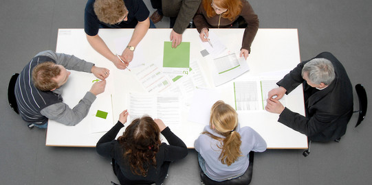 Studierende und Dozierende sitzen gemeinsam an einem Tisch und arbeiten.