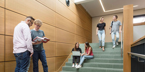 Zwei Studierende laufen eine Treppe herunter, zwei sitzen auf der Treppe und zwei stehen davor.